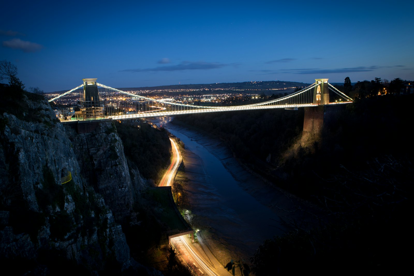 A lit suspension bridge spanning a river gorge at dusk, city lights in the distance