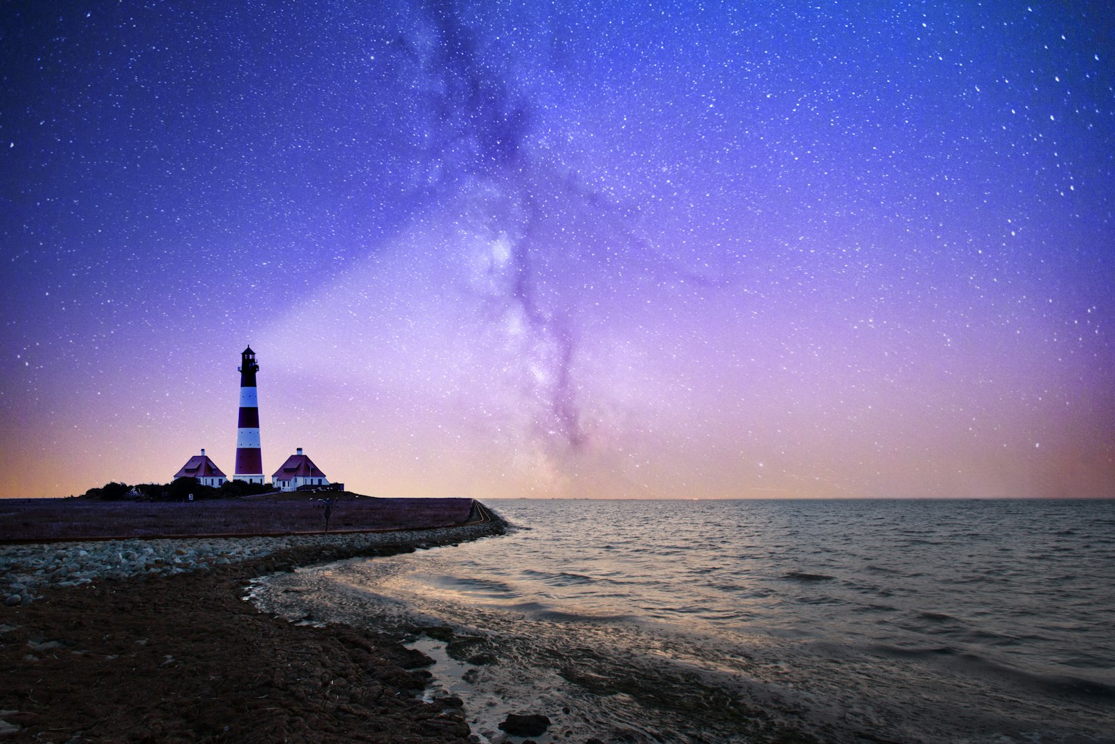 A striped coastal lighthouse beneath the Milky Way, its beam sweeping across a starry night sky